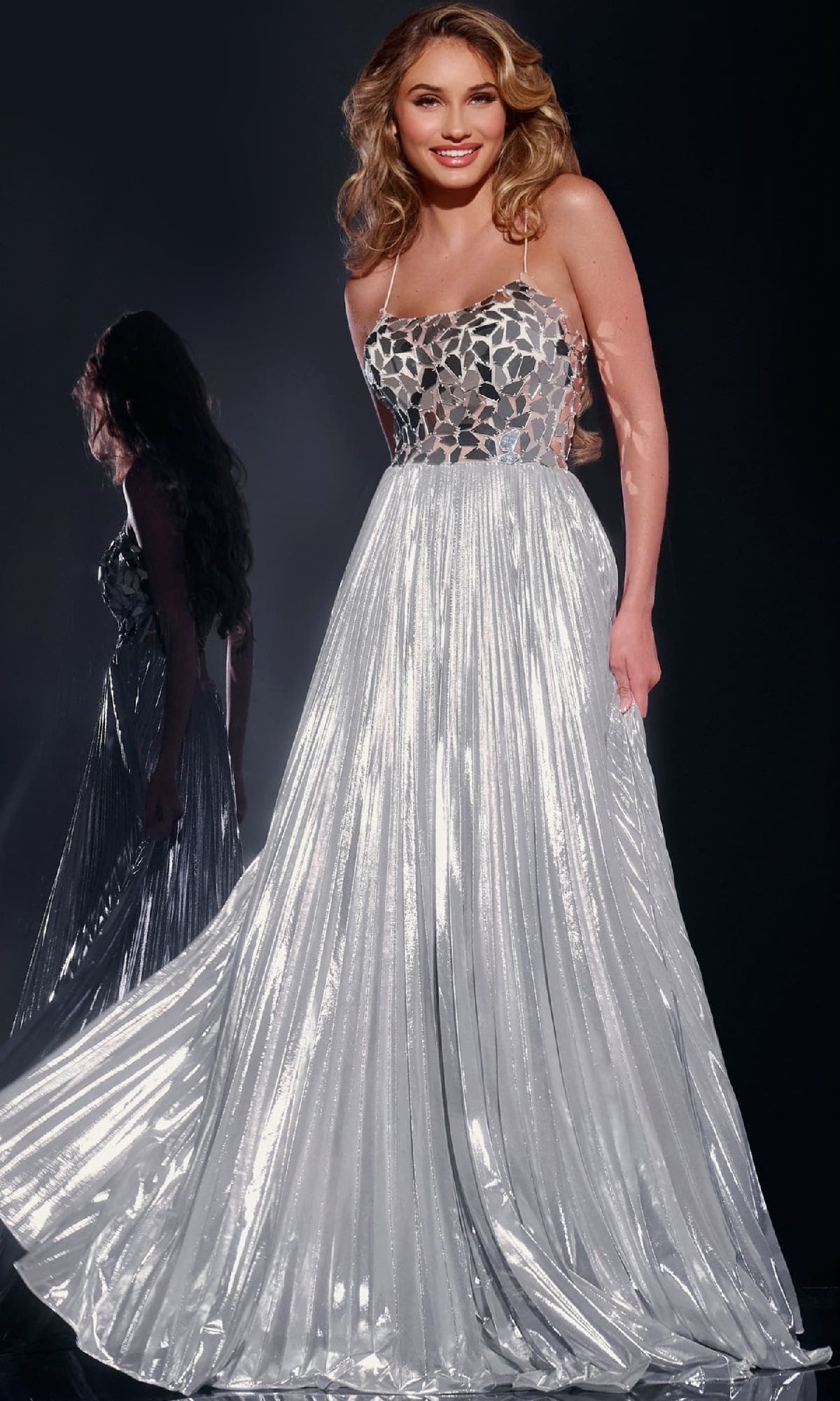 A woman wears the Jovani 41117, a floor-length silver pleated A-line prom dress with a metallic skirt and a fitted bodice featuring a geometric pattern.